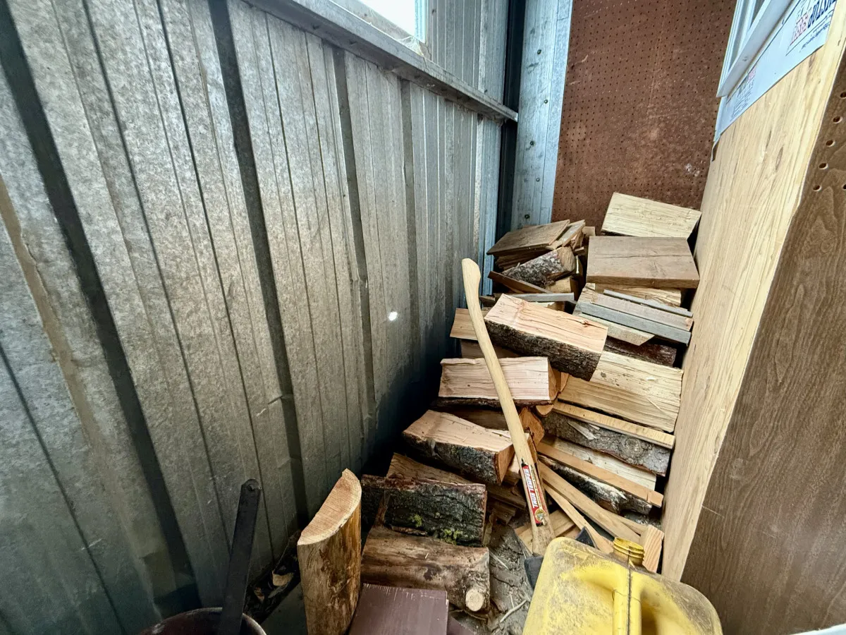 A cozy, rustic stack of firewood with a wooden splitting axe and metal container set against a wooden wall. The image exudes warmth, preparation, and the satisfaction of a wood-burning fire after a long day in the snow.