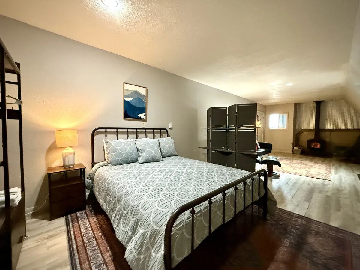 The image showcases a cozy interior featuring a plush queen bed, a softly glowing nightstand lamp, inviting wall art, and a classic wood stove radiating warmth. The soft morning light filters through the window, enhancing the intimate, relaxing atmosphere designed for unwinding after adventurous days.