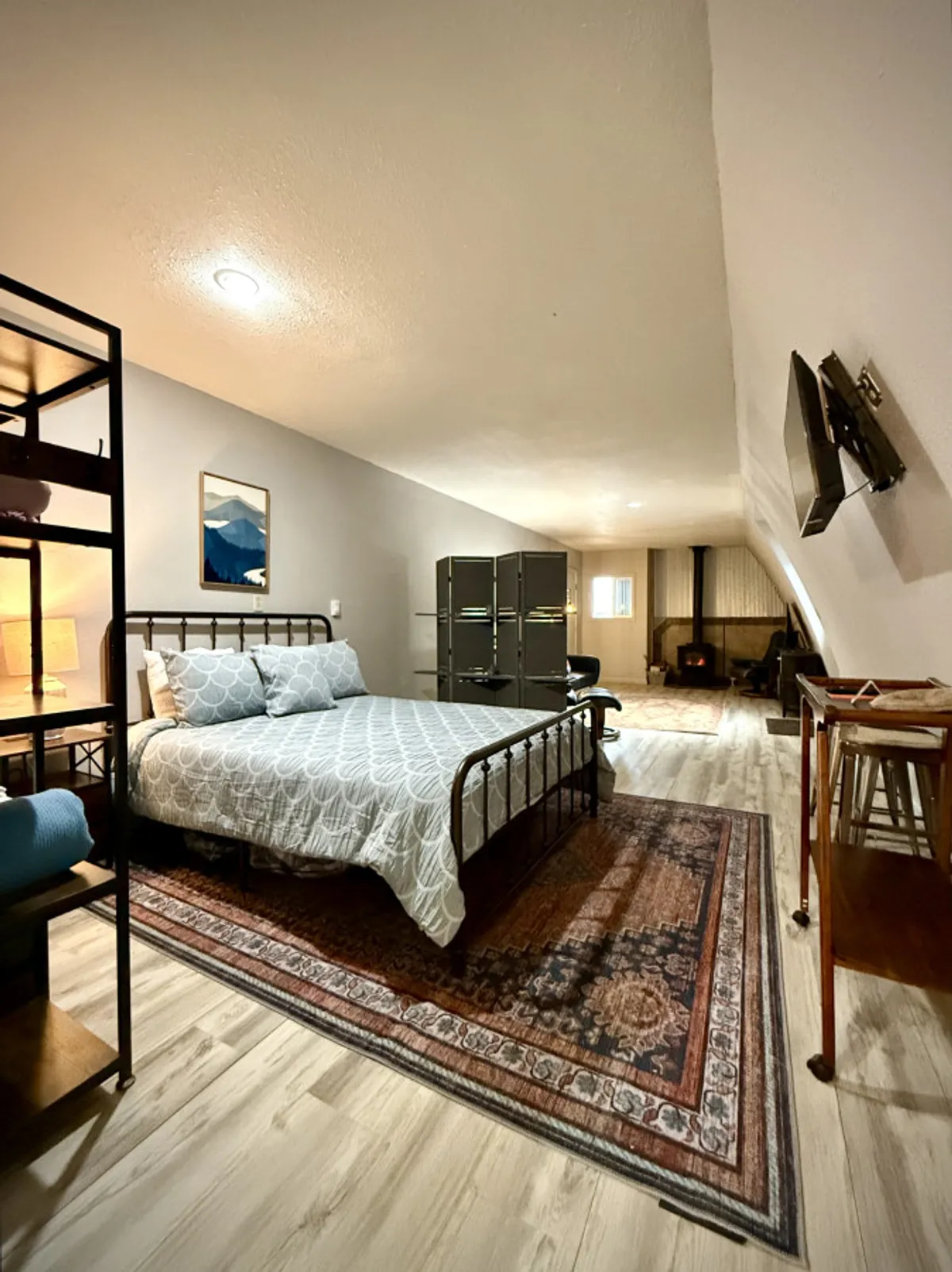 This interior image features a plush queen bed with inviting linens, a cozy fireplace on one side, a decorative shelf, and artwork adorning the wall. The ambient glow from the fire suggests warmth and relaxation, creating the perfect setting for peaceful nights and restful mornings.