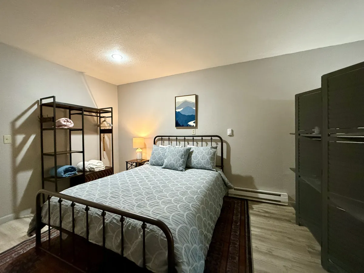 A bright, comfortable bedroom scene showcasing a well-made bed, a nightstand with a lamp, and tasteful artwork above the headboard. Open shelving adds a touch of personal style, and natural light filters into the space, enhancing the sense of peace and privacy.