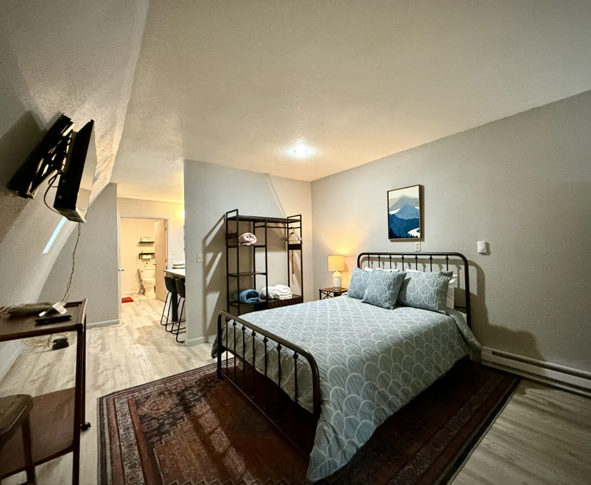 A welcoming bedroom scene with a bed dressed in patterned linens, a shelf with neatly folded clothes, a framed mountain painting, and a TV mounted on the wall. The space radiates comfort and invites guests to unwind and relax after a day of adventure.