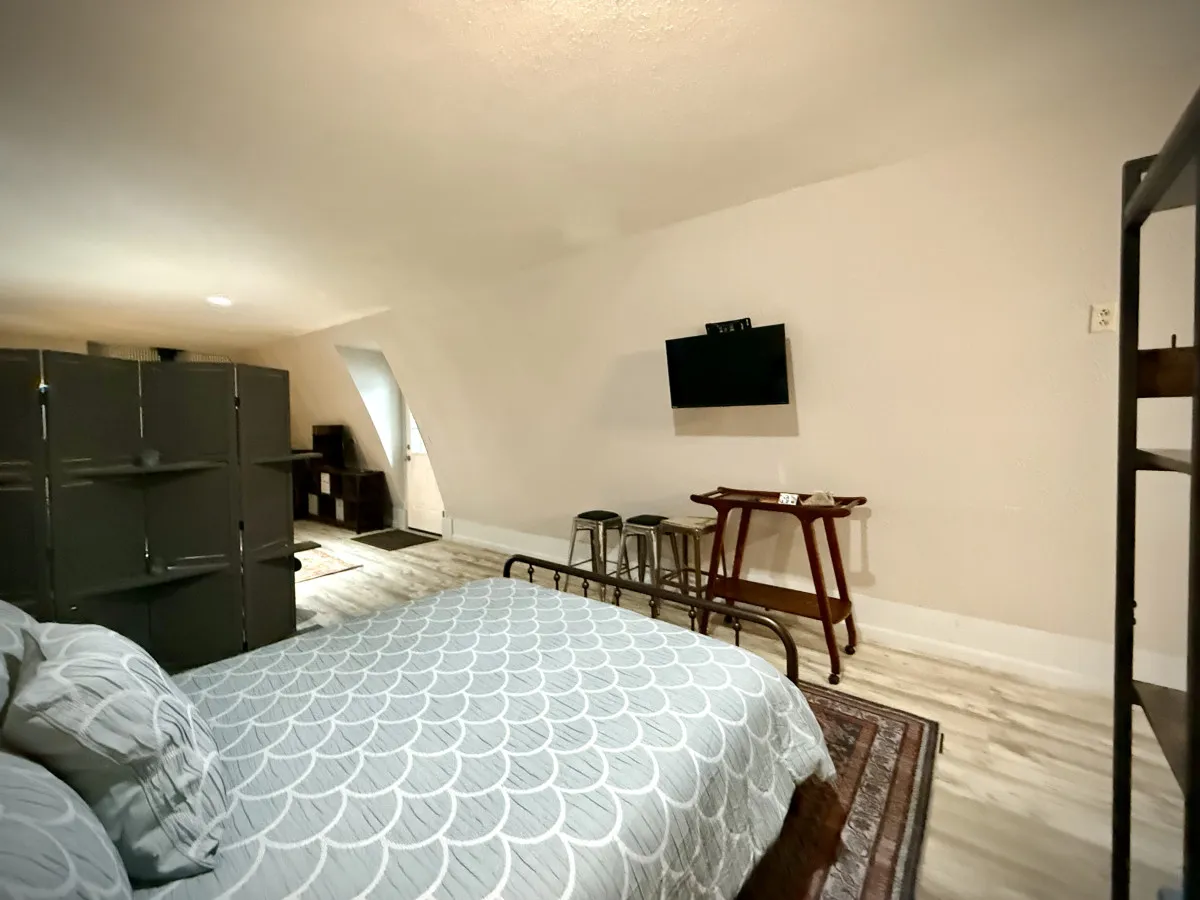 This image features a studio interior with a bed adorned in patterned bedding, a small table with stools perfect for sharing information or planning outings, and a television mounted on the wall. A room divider subtly separates spaces, evoking both coziness and functionality.