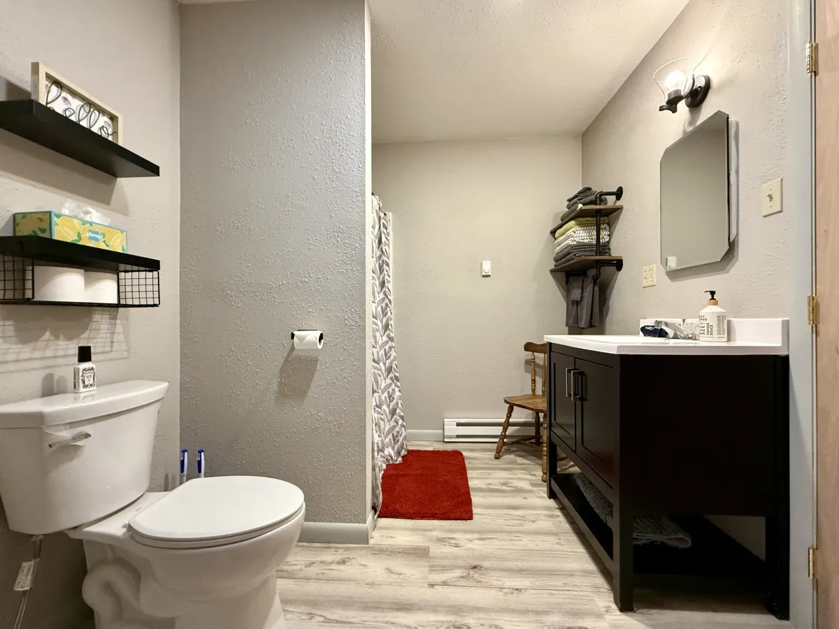 A calm and organized full bathroom complete with a toilet, a vanity with a sink, a shower area, and neatly folded towels on a shelf. The design offers a refreshing and soothing environment for morning or evening routines.