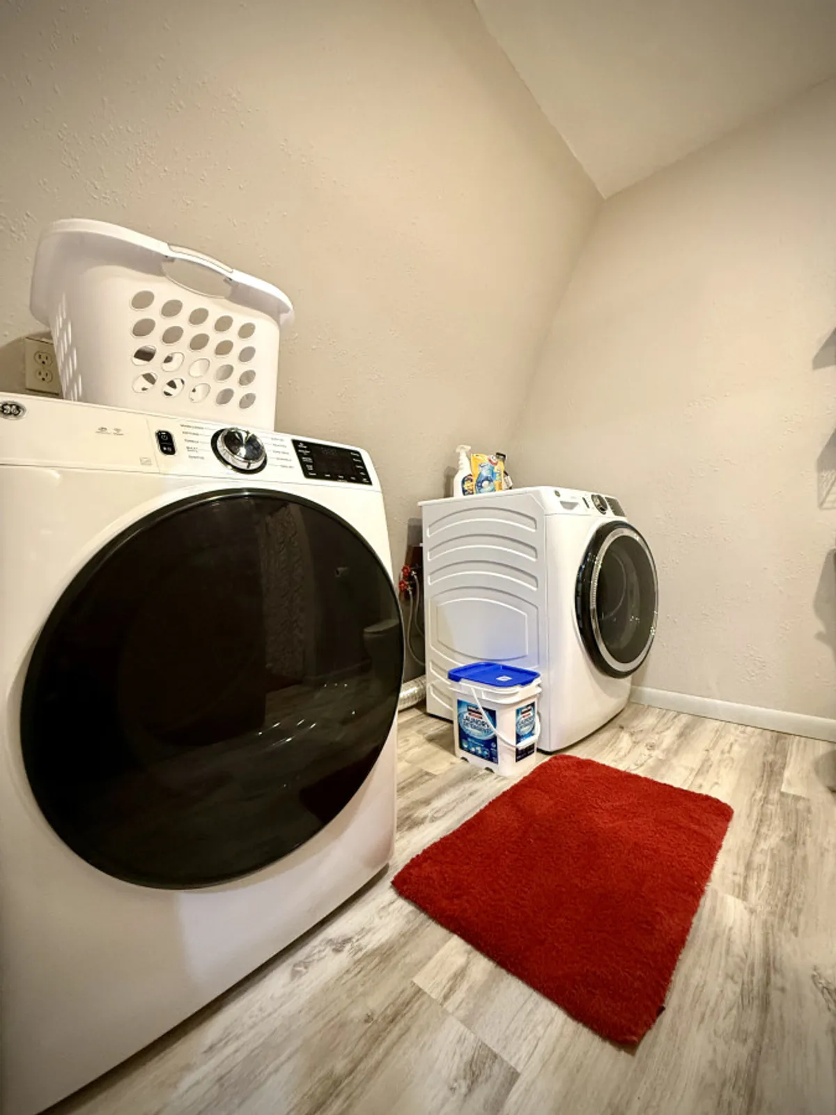 A bright and tidy laundry area featuring a washing machine and dryer side by side, complemented by a laundry basket and a cozy red rug. The space is organized and practical, offering a welcoming spot for keeping clothes fresh during any stay.