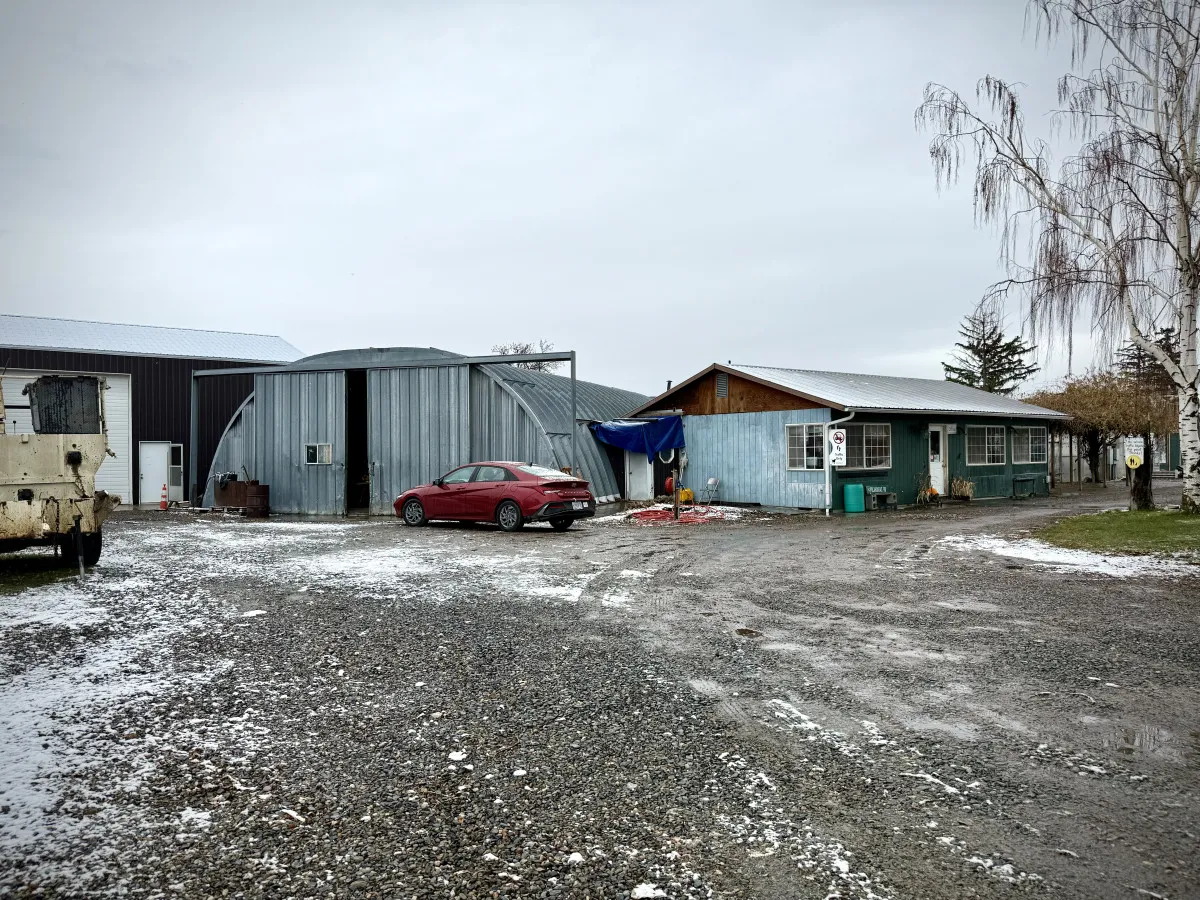 An exterior shot of a property surrounded by snowy ground, a red car parked nearby, a metal building, and a wooden house. The setting feels welcoming and practical, suggesting easy access to outdoor adventures and a warm homecoming.